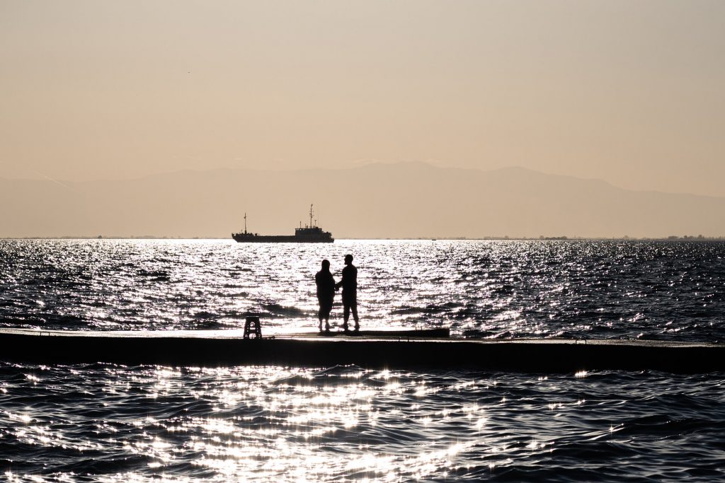Ein Paar steht Hand in Hand vor dem glitzernden Meer, ein Schiff im Hintergrund.