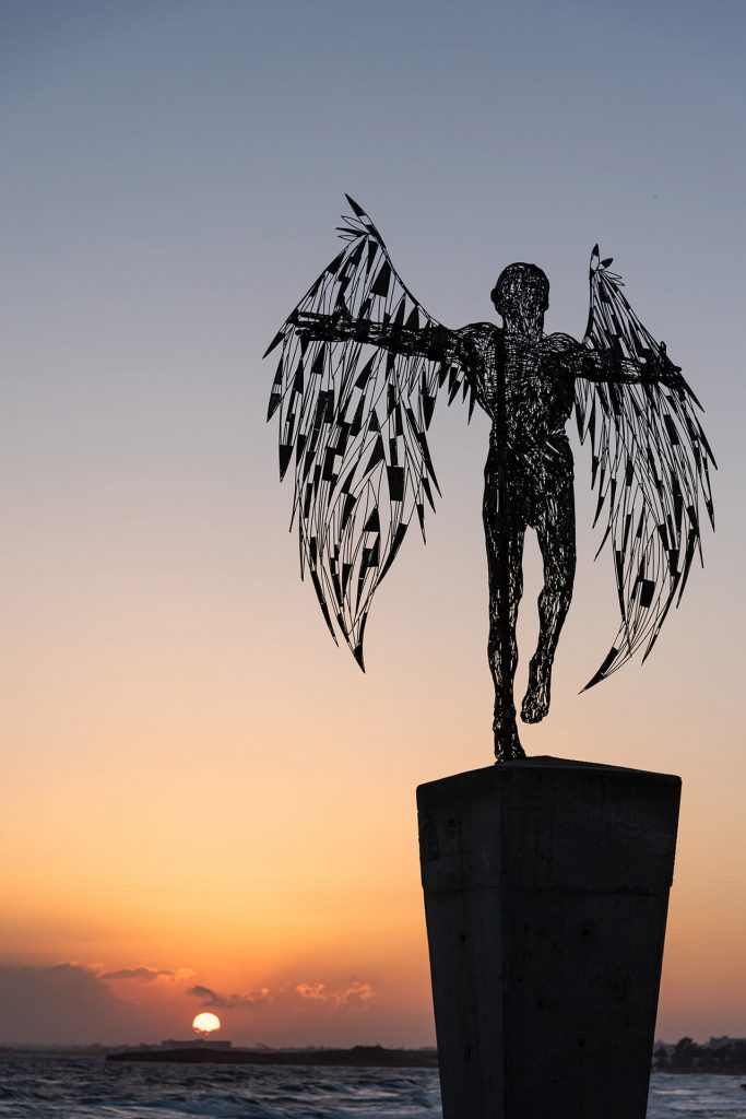 Skulptur von Ikarus mit Flügeln bei Sonnenuntergang am Meer