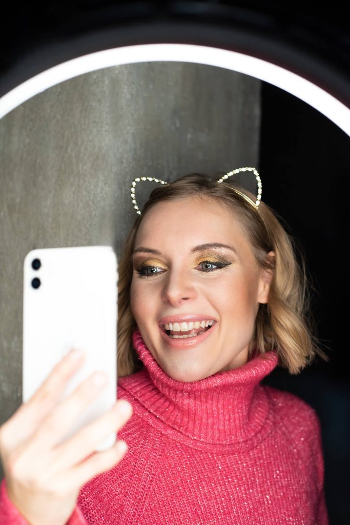 Schauspielerin Carolin Lehmann lacht herzlich, blickt ins Handy, pinker Pulli und funkelnde Katzenohren sichtbar.