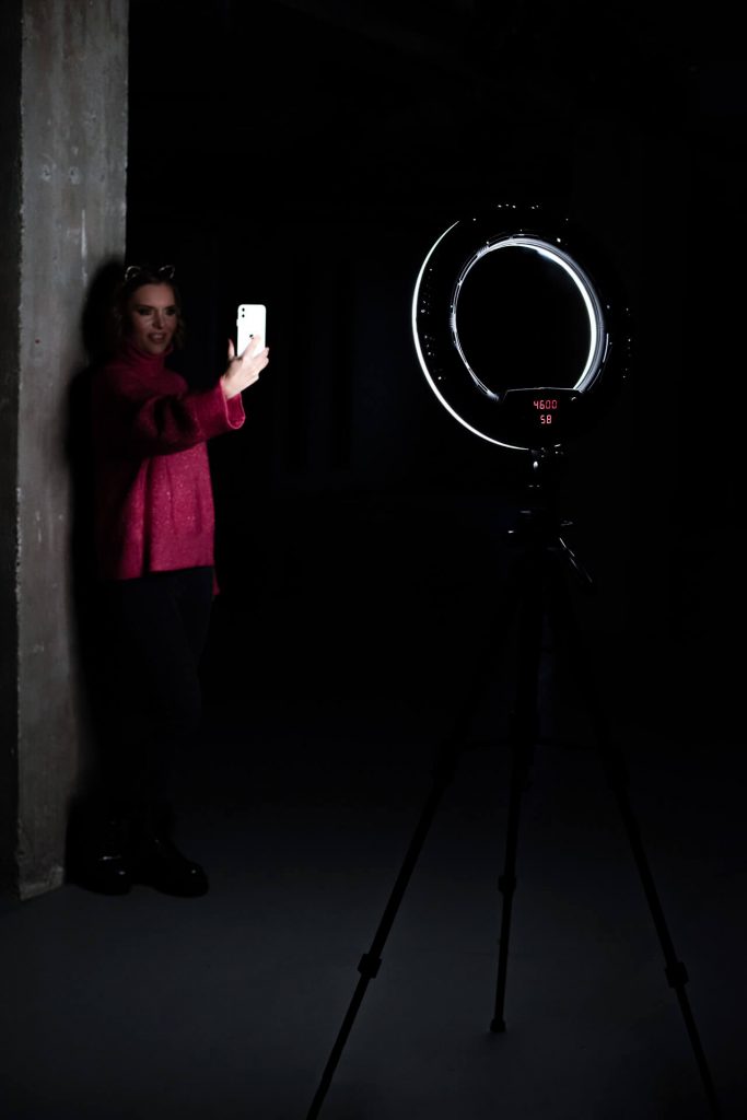 Schauspielerin lehnt an einer Betonwand und nimmt sich mit einem Smartphone vor einem Ringlicht auf – Behind-the-Scenes der Theaterproduktion Lehmann.