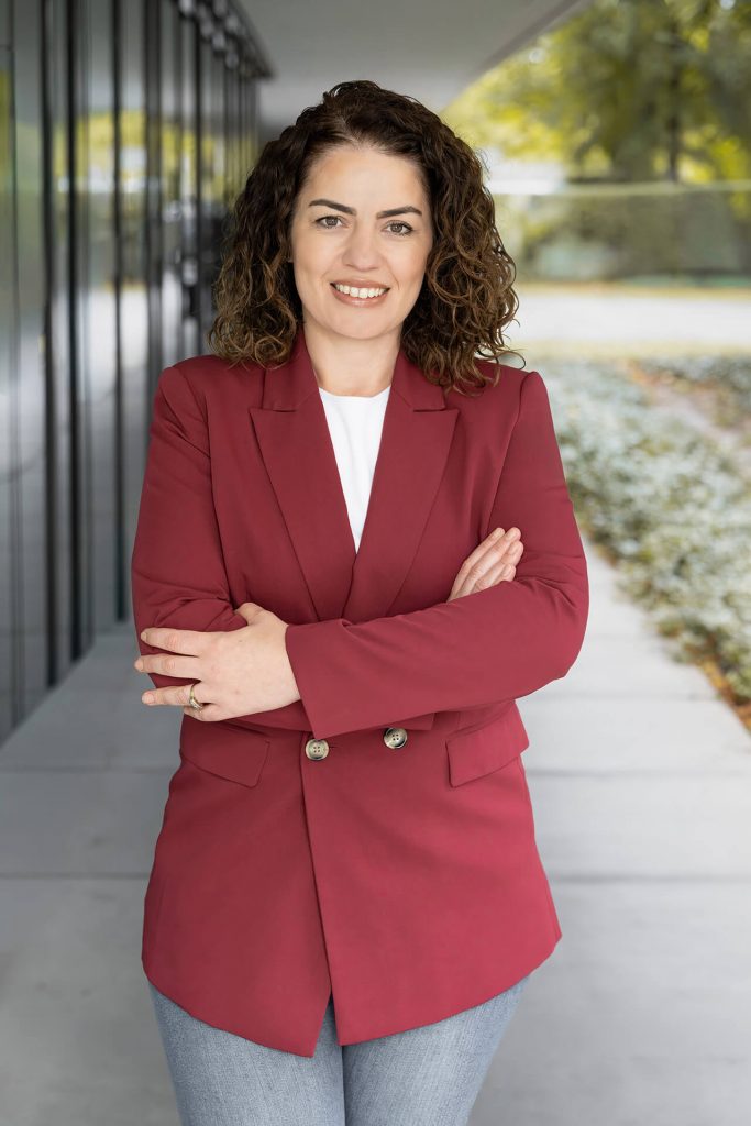 Authentisches Bewerbungsfoto einer selbstbewussten Frau im Business-Outfit mit freundlichem Lächeln im Außenbereich.