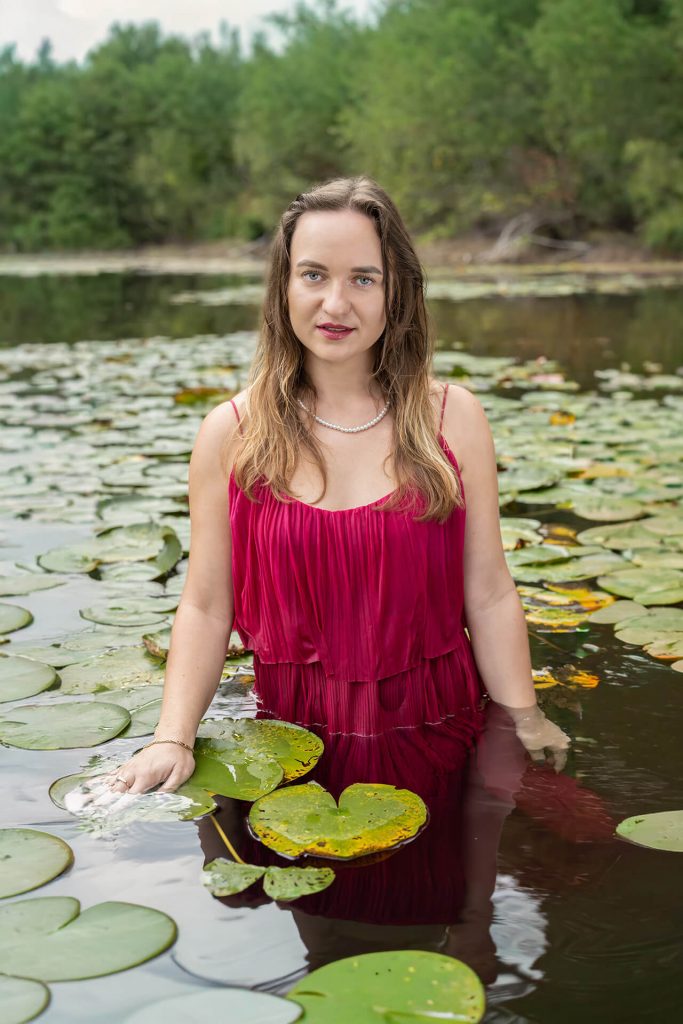 Frau in elegantem roten Kleid steht im Wasser zwischen Seerosen, yourphotoandstory