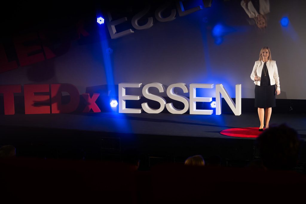 Rednerin auf der Bühne bei TEDxEssen mit Lichtreflexionen auf dem TEDx-Schriftzug, Mikaela Gallon mit der Rede: Mind the Gap: A step by step guide for impactful presence