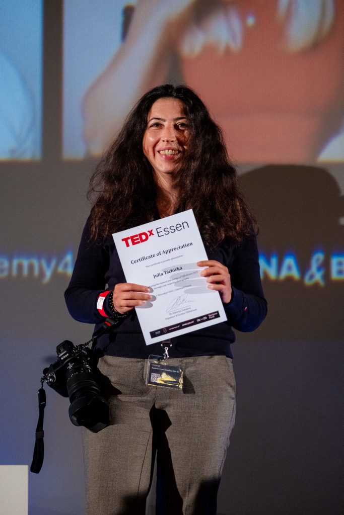 Fotografin hält TEDxEssen-Zertifikat auf der Bühne, warmes Bühnenlicht und Kamera sichtbar.