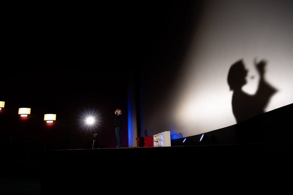 Sprecherin auf einer Bühne mit TEDx-Branding, dramatischer Schattenprojektion und stimmungsvoller Bühnenbeleuchtung, eingefangen bei einem Live-Event.
