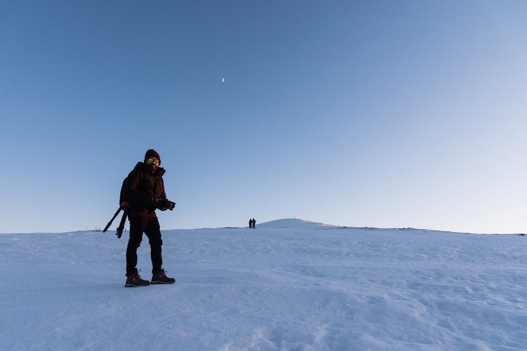 Eine Person mit Kamera steht in einer weiten Schneelandschaft unter klarem Himmel mit Mond.