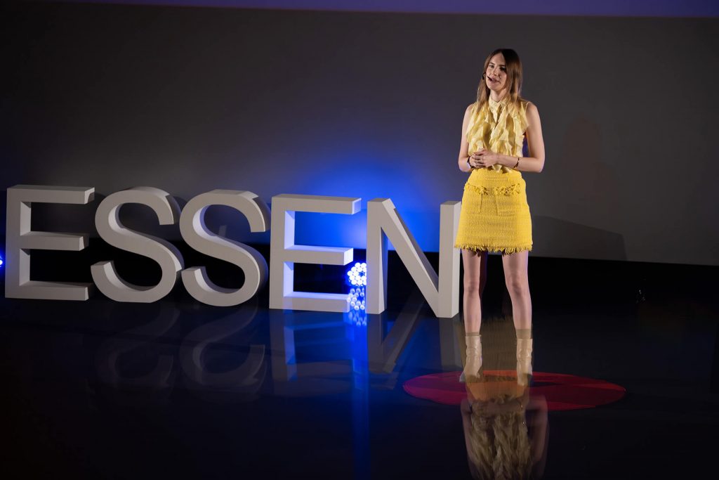 Junge Rednerin im gelben Kleid mit Reflektion auf der TEDx-Bühne, Céleste Spahić mit der Rede: Does ChatGPT Make Us Dumb or Finally Human?