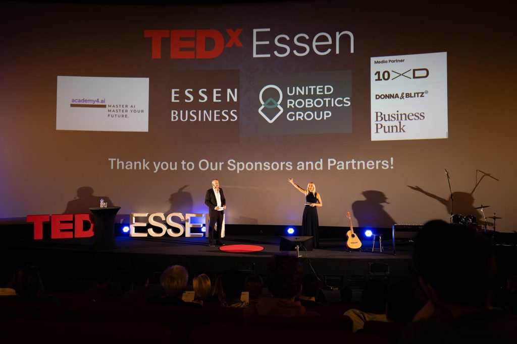 Sponsorenwand auf Leinwand bei TEDxEssen mit Moderatoren Dirk Brueckner & Dr. Viktoriya Sokolova auf der Bühne