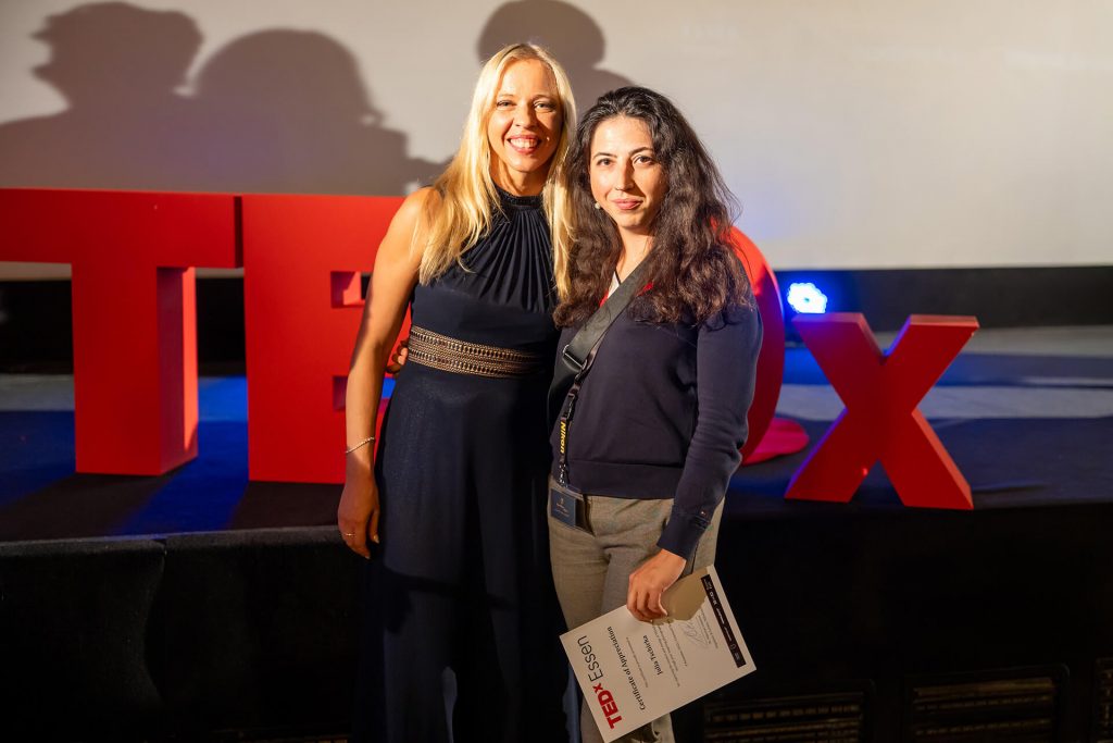 Dr. Viktoriya Solova und Dr. Julia Tschirka stehen vor dem roten TEDx-Schriftzug in Essen und lächeln in die Kamera.