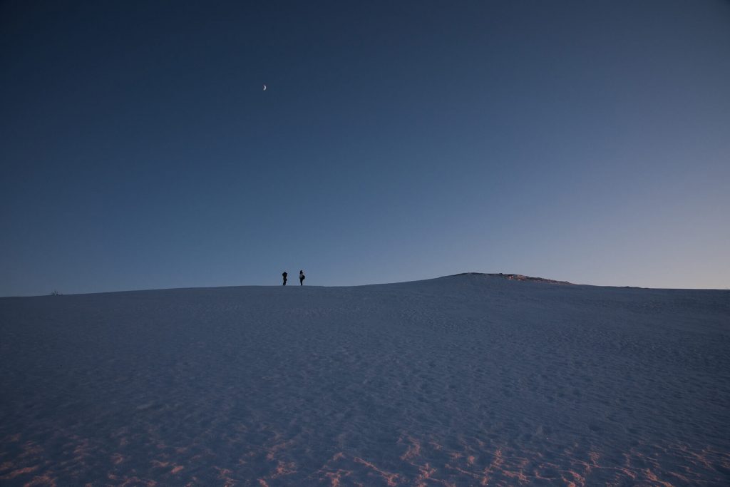 Zwei Personen stehen auf einer weiten, verschneiten Fläche unter einem klaren Winterhimmel mit Mond.
