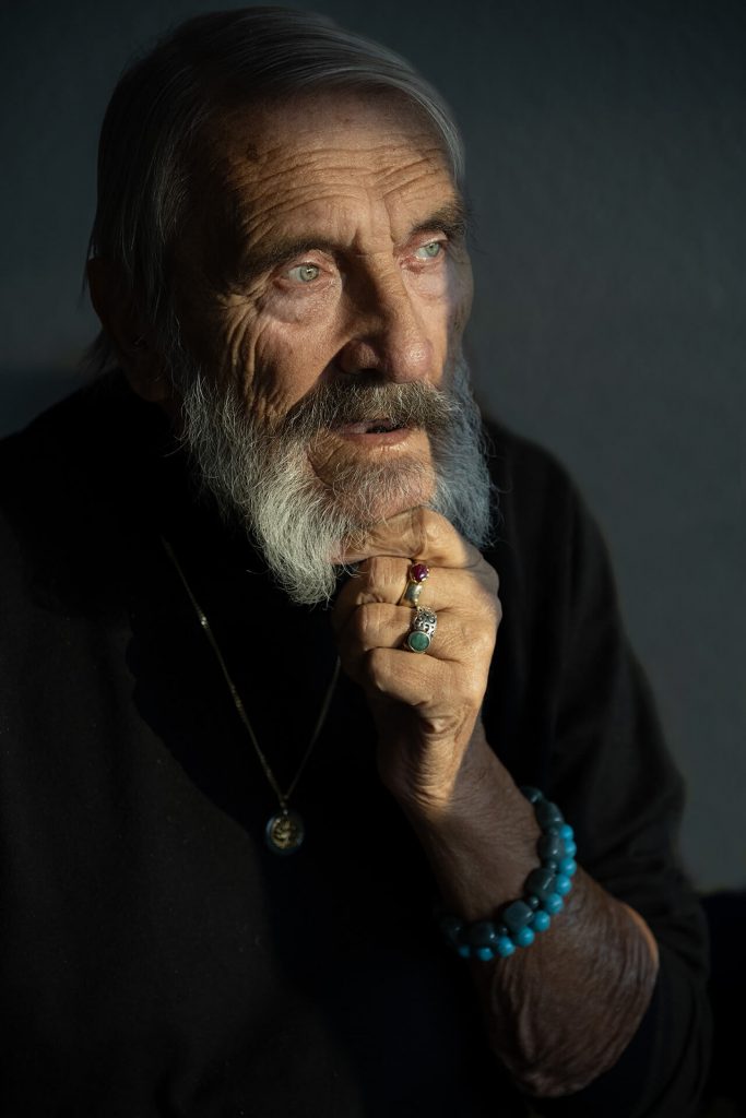 Älterer Mann mit grauem Bart und Schmuck, im Seitenlicht fotografiert, ruhiger Blick vor dunklem Hintergrund