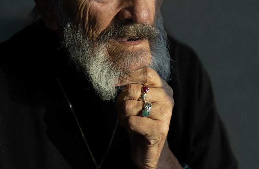 Älterer Mann mit grauem Bart und Schmuck, im Seitenlicht fotografiert, ruhiger Blick vor dunklem Hintergrund