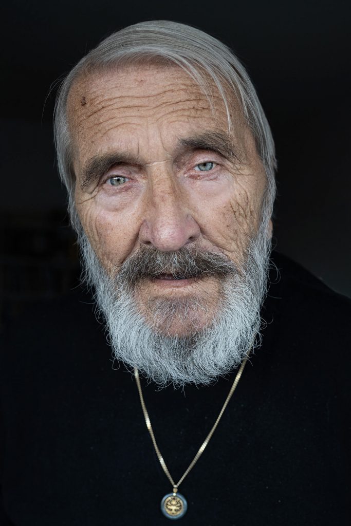 Frontalporträt eines älteren Mannes mit grauem Bart, direkter Blick in die Kamera, dunkler Hintergrund