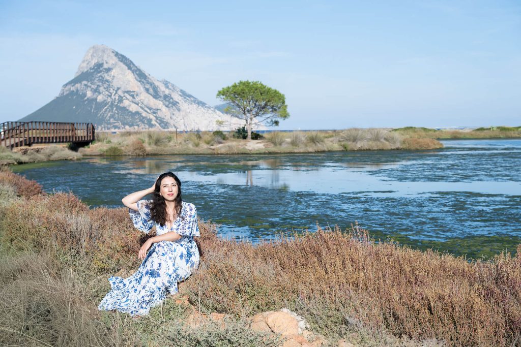 Julia Tschirka im weiß-blauen Kleid am Ufer einer Lagune auf Sardinien, im Hintergrund die Felseninsel Tavolara.